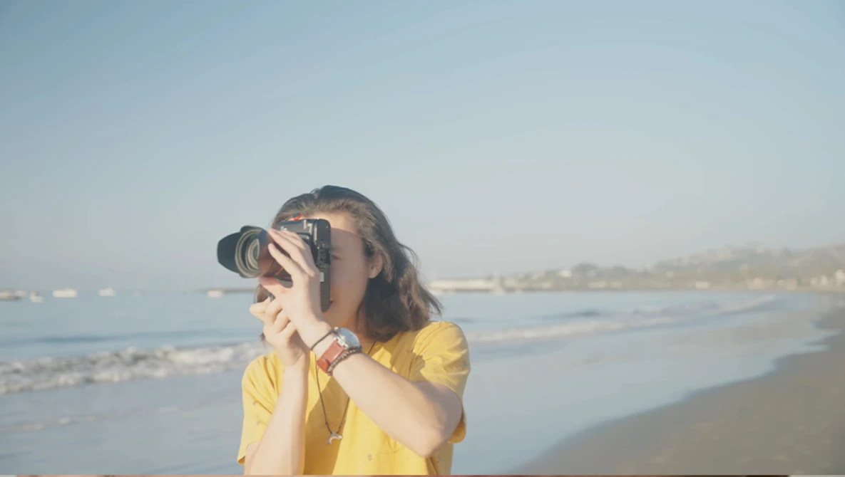 Woman in yellow shirt holding camera up to her face on a beach at golden hour.