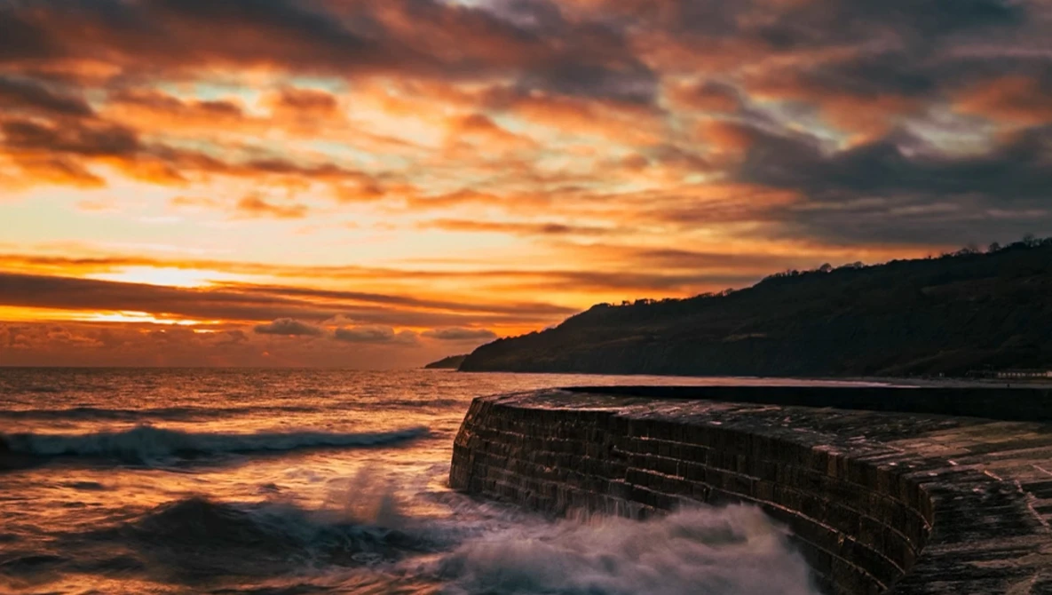 Dramatic coastal sunset with layered rock formations in foreground and mountainous headland silhouetted against golden sky.