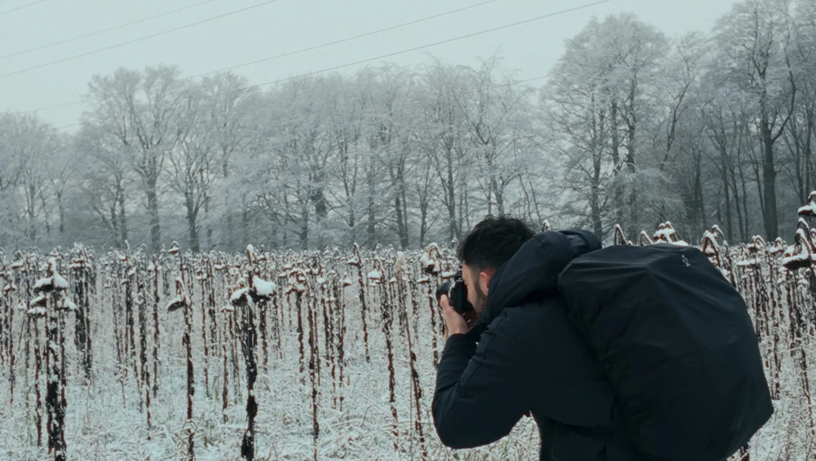 Photographer in black jacket shooting a frost-covered cornfield with bare trees in misty background.