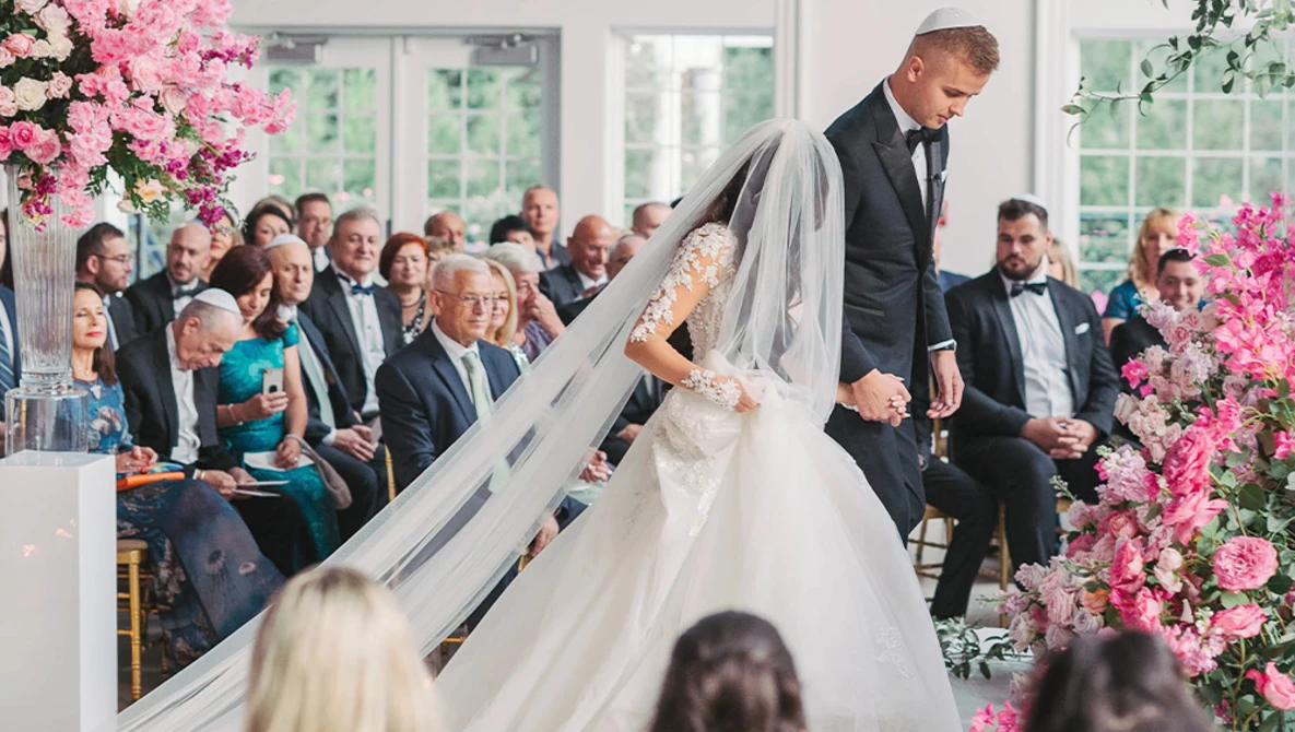 Groom helping bride with her veil during wedding ceremony indoors with pink floral arrangements.