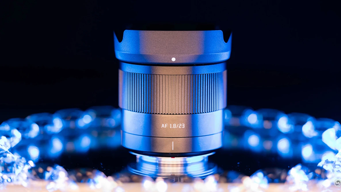 Close-up of a blue and bronze lens with AF 1.8/23 marking, surrounded by bokeh lights on dark background.