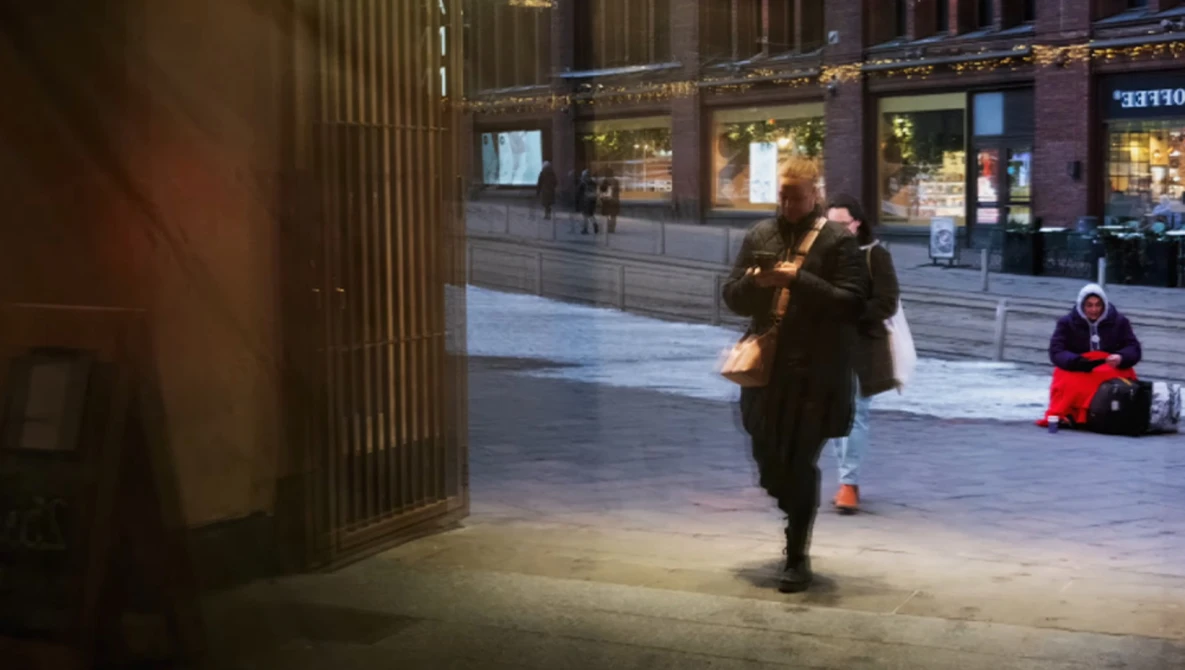 Woman in dark coat walking across an urban plaza at dusk, storefront windows visible in background.