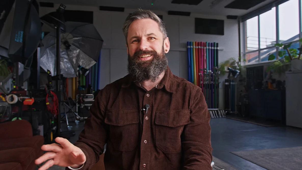 Bearded man in brown shirt gesturing while speaking in a photography studio with equipment and large windows.