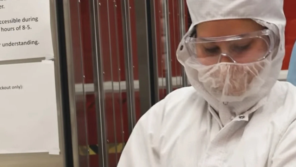 Person wearing full white protective gear including hood, face shield, and goggles in an indoor setting.
