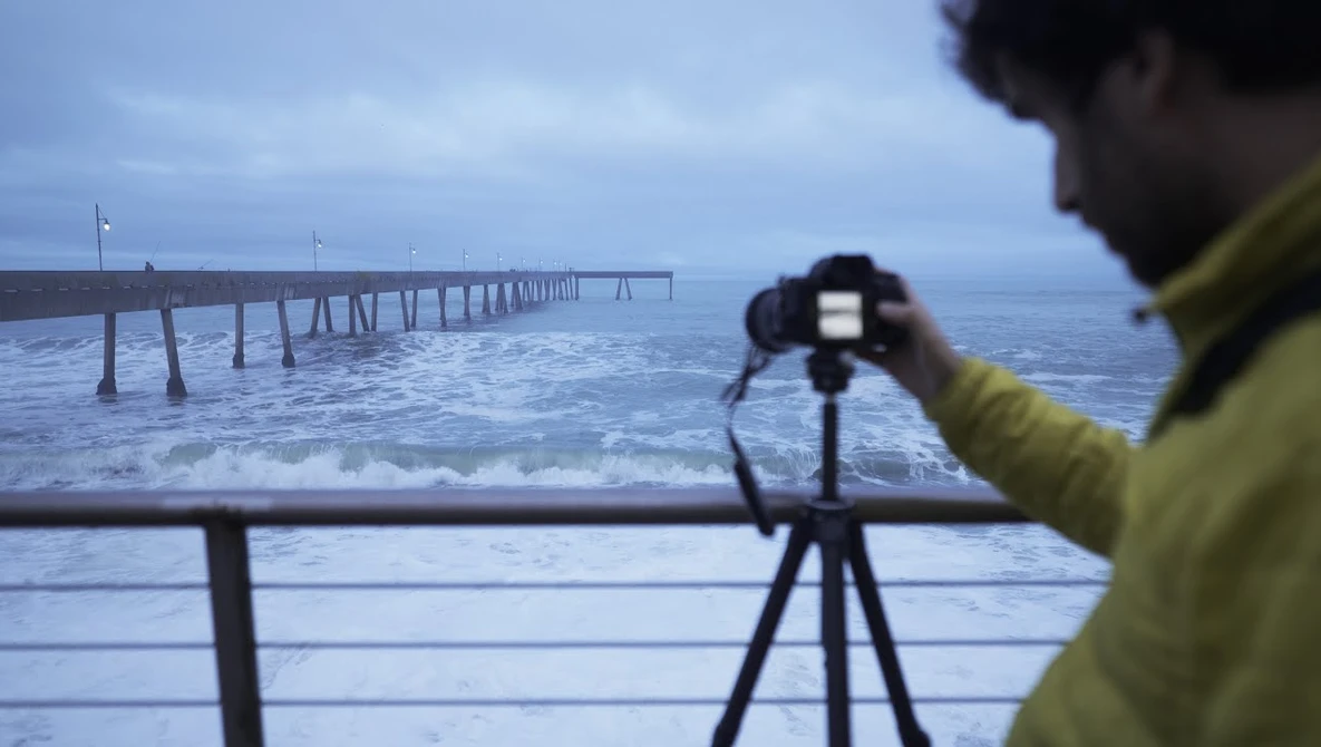 Photographer in yellow jacket capturing pier extending into ocean at twilight using tripod-mounted camera.