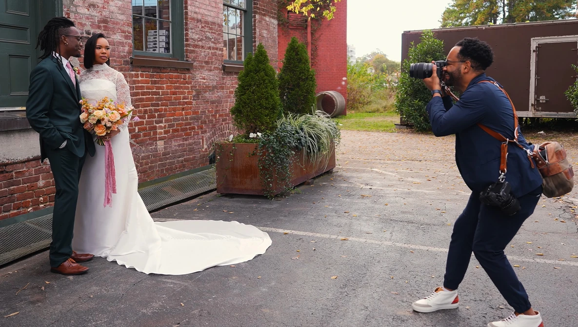 Wedding photographer capturing bride and groom posing beside a brick building during an outdoor portrait session.