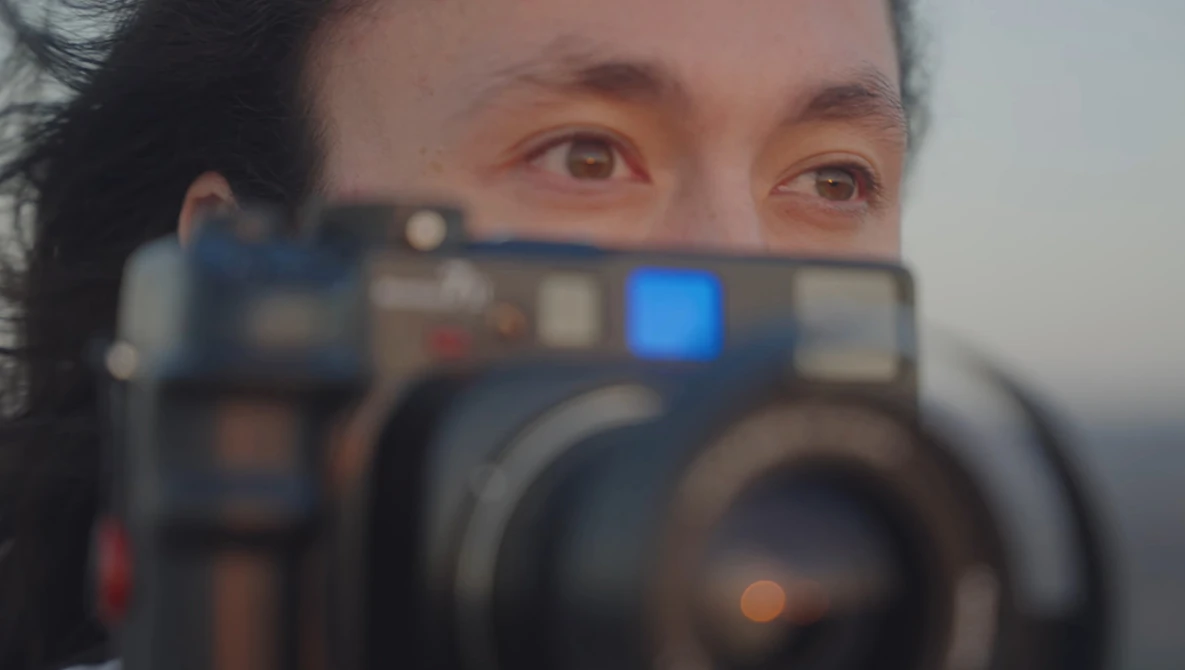 Photographer holding a professional video camera with blue display screen visible, shot through the camera lens with shallow depth of field.