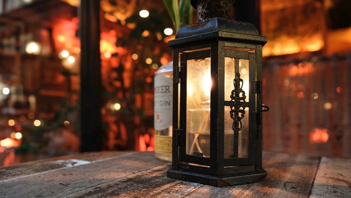 Black metal lantern with illuminated interior sitting on wooden surface with warm bokeh lights blurred in background.