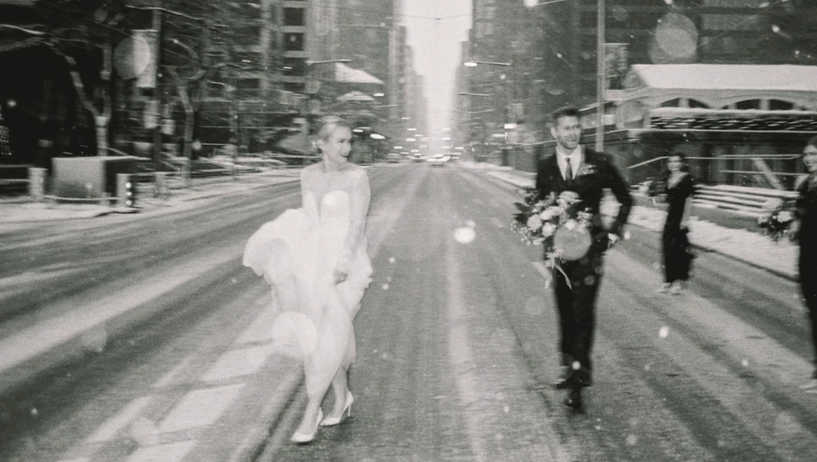 Bride in white dress running down urban street with groom and wedding party following behind.