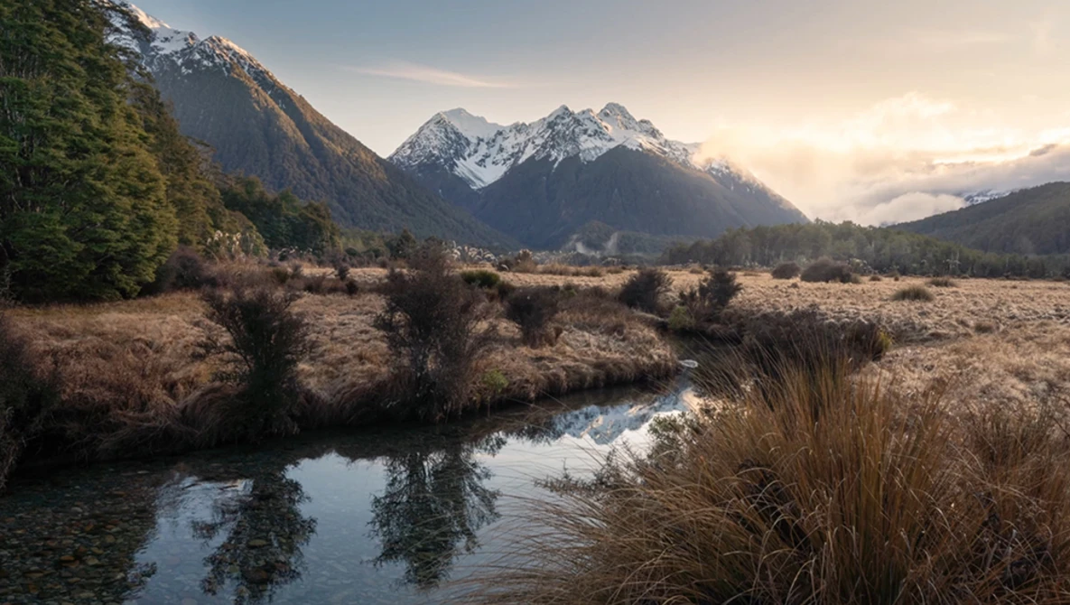 Snow-capped mountain peaks rise above a meandering stream in an expansive valley at golden hour.