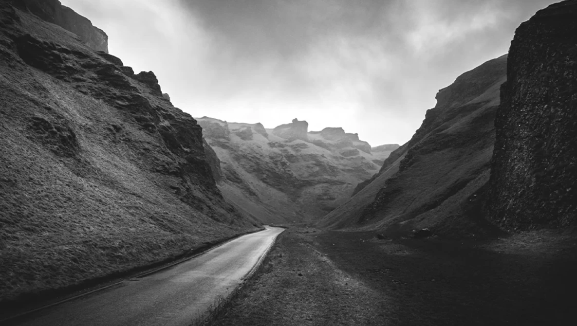 Narrow asphalt road cutting through a volcanic canyon with steep dark rock walls under an overcast sky.