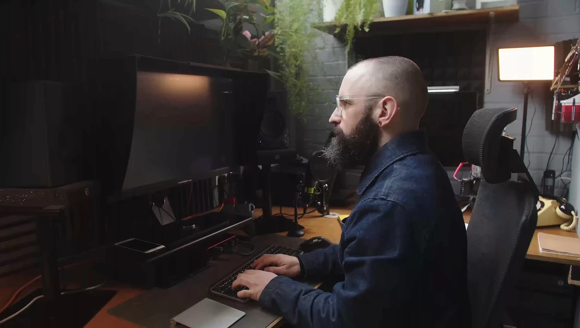 Bearded man at desk working on computer in a dark creative studio space.