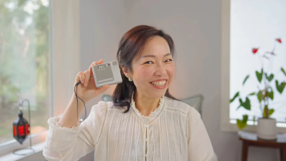 Woman holding a compact digital camera toward the camera with a bright smile indoors.