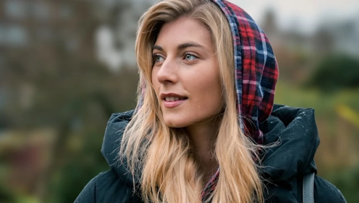 Woman wearing a dark jacket and red plaid hood, photographed outdoors during overcast daylight.
