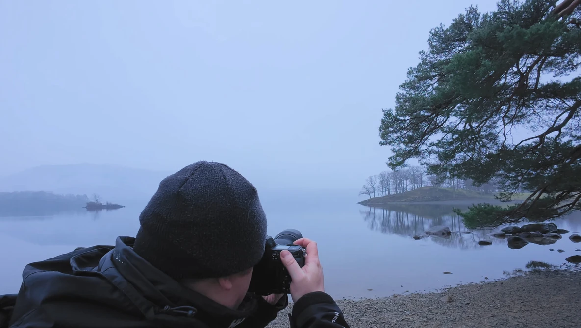 Photographer shooting landscape at dawn by a misty lake with pine trees reflected in still water.