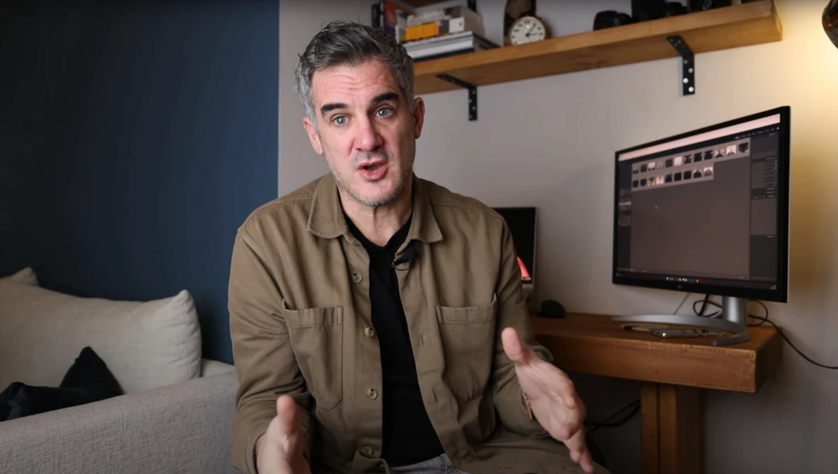 Man in tan jacket speaking to camera in home studio with monitor displaying editing software.