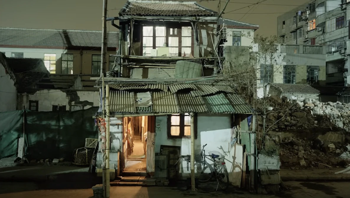 Weathered two-story residential building with illuminated doorway at night, surrounded by older structures.