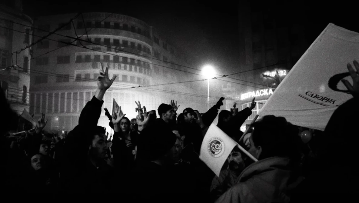 Nighttime crowd protest with raised hands and flags, urban buildings lit by street lamps.