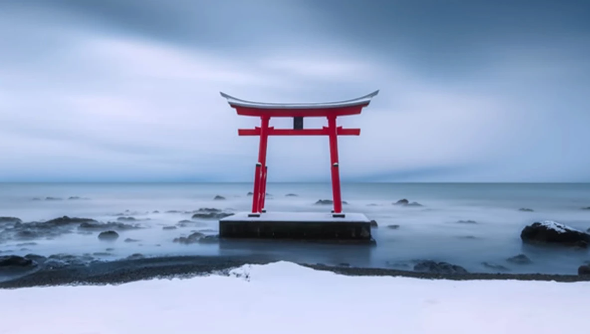 Red torii gate standing in shallow ocean water with snow-covered beach and rocky coastline.