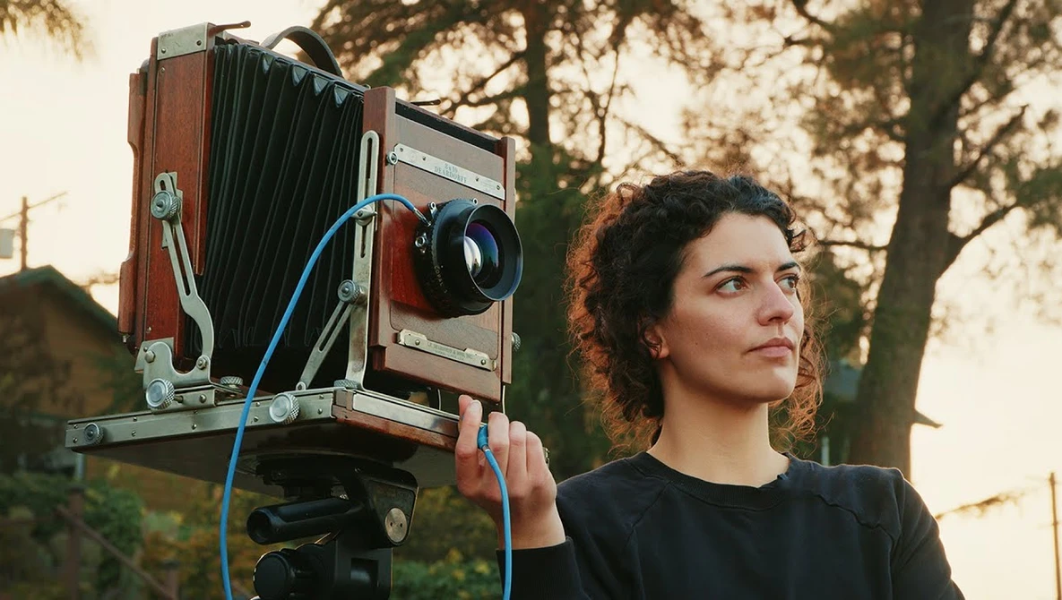 Photographer holding a large format camera with blue tubing attached, outdoors during golden hour.