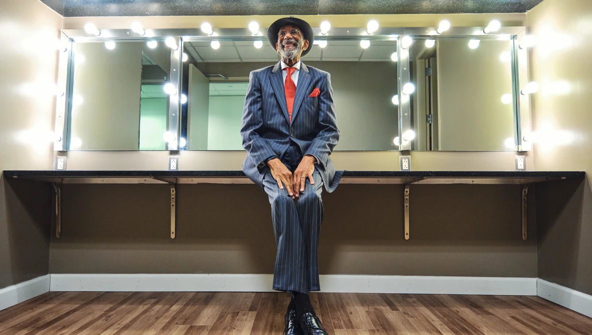 Person in blue pinstriped suit and red tie sitting on vanity counter beneath illuminated mirror with multiple bulbs.