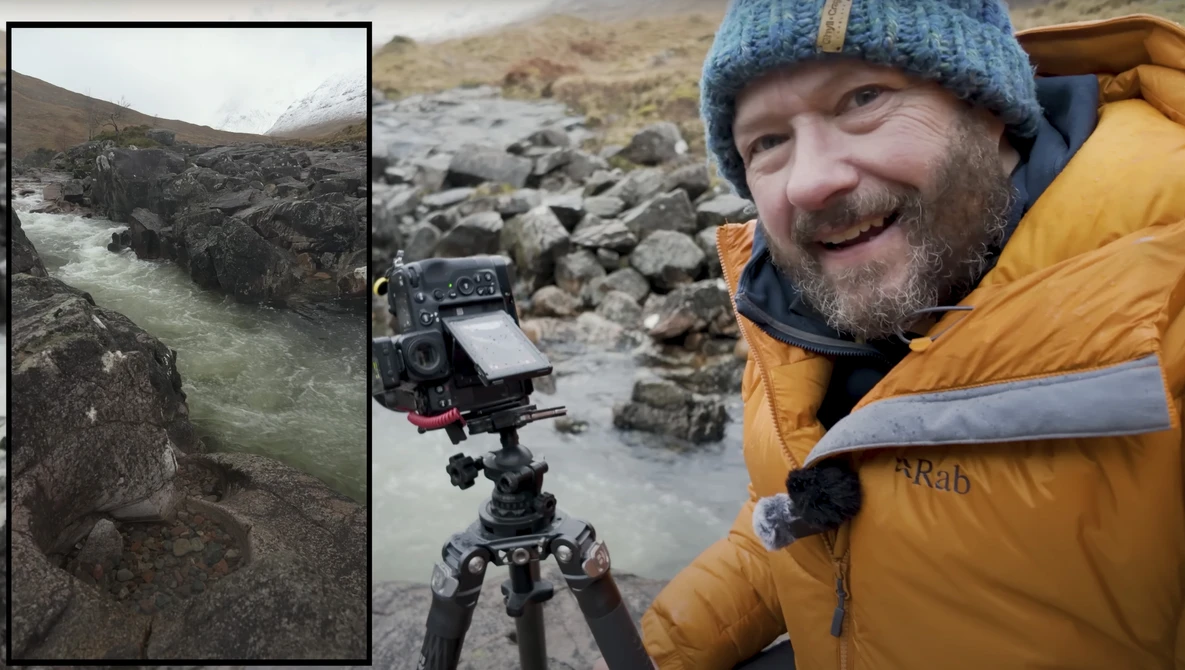 Filmmaker smiling at camera while operating cinema camera on tripod beside rushing glacial river in rocky mountain valley.