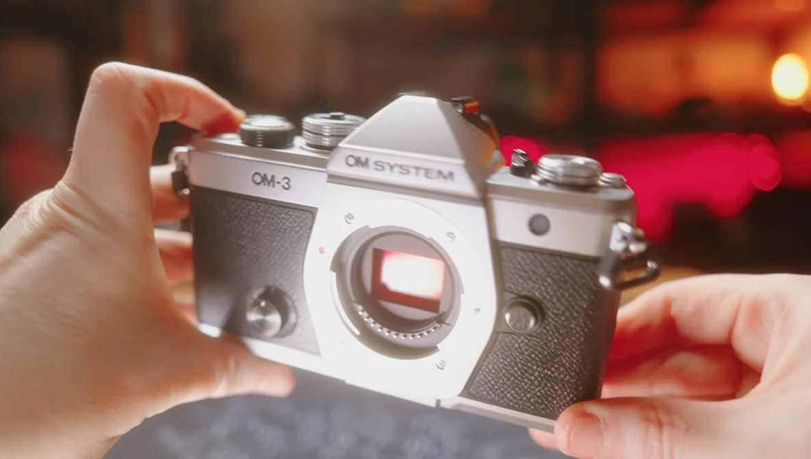 OM System OM-3 film camera held in hands against warm-toned blurred background.