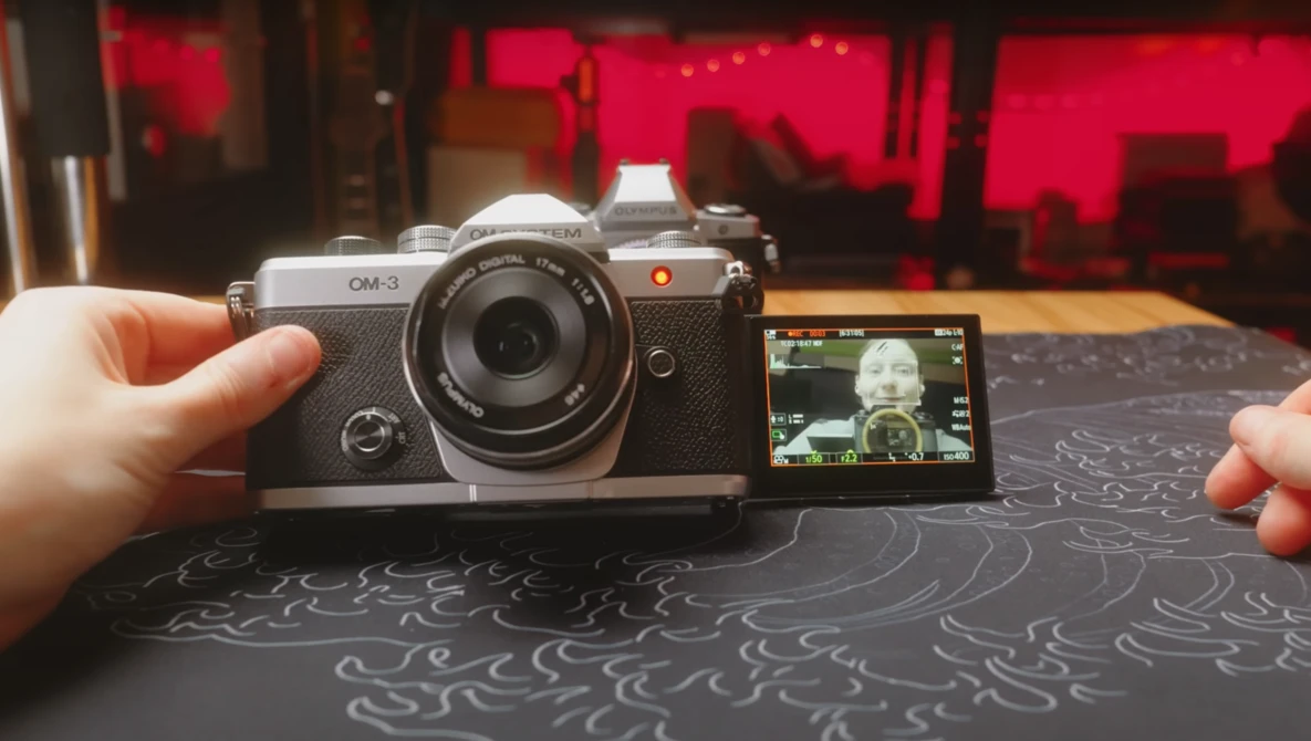 Hands holding a compact rangefinder camera with articulating rear screen displaying a portrait.