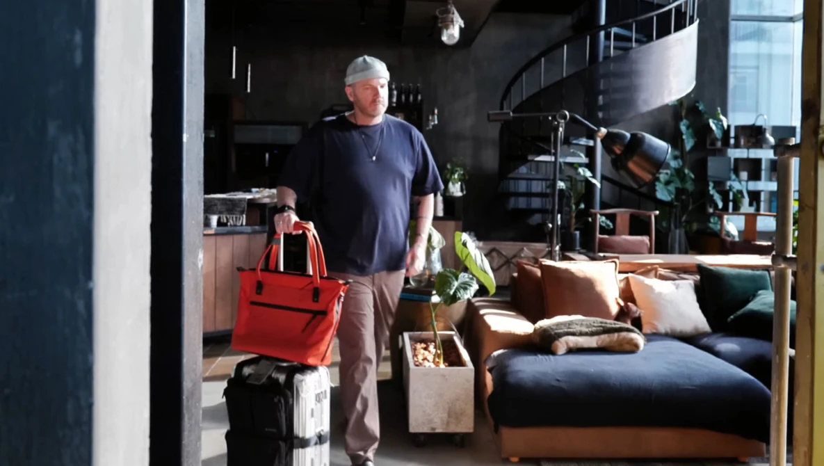Man standing in a loft studio space holding a red bag next to packed luggage and moving boxes.
