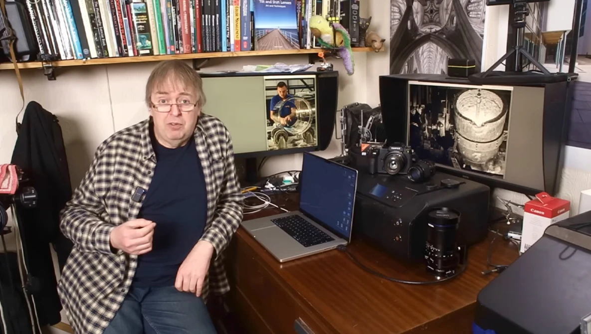 Photographer in home studio workspace with laptop, camera equipment, and shelves of books and photography materials.