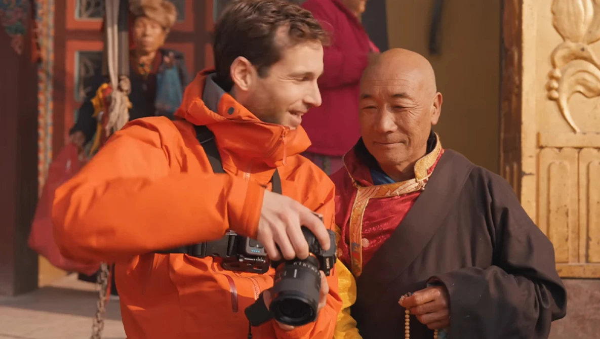 Man in orange jacket holding binoculars speaks with elderly man wearing brown robe and prayer beads.
