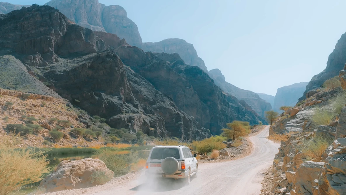 White SUV driving on a dusty mountain road through a dramatic canyon with steep rocky cliffs.