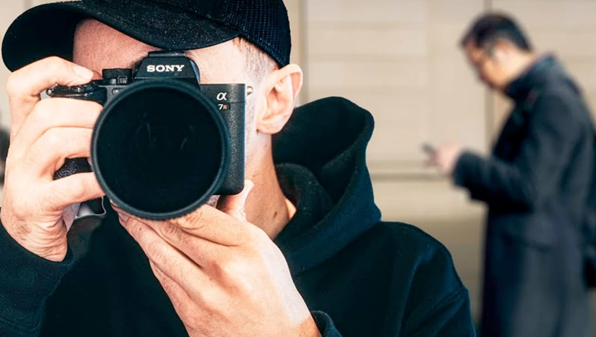 Photographer holding Sony mirrorless camera up to face while wearing black cap and jacket.