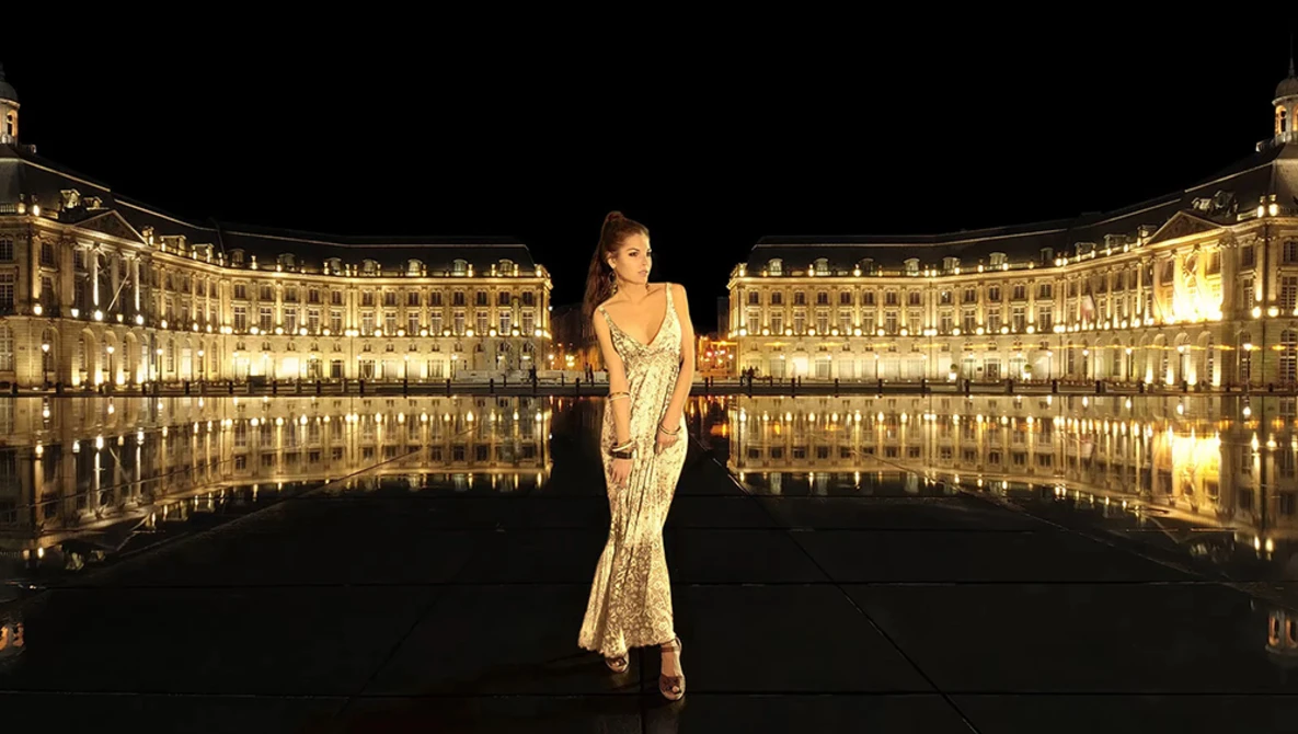 Woman in an elegant beaded gown standing centered in a symmetrical plaza reflected in still water at night.