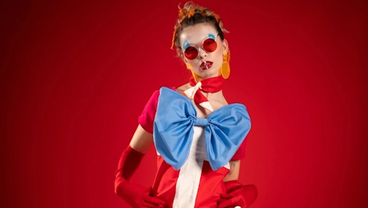 Woman in red jacket and blue bow tie against vibrant red background wearing round sunglasses.