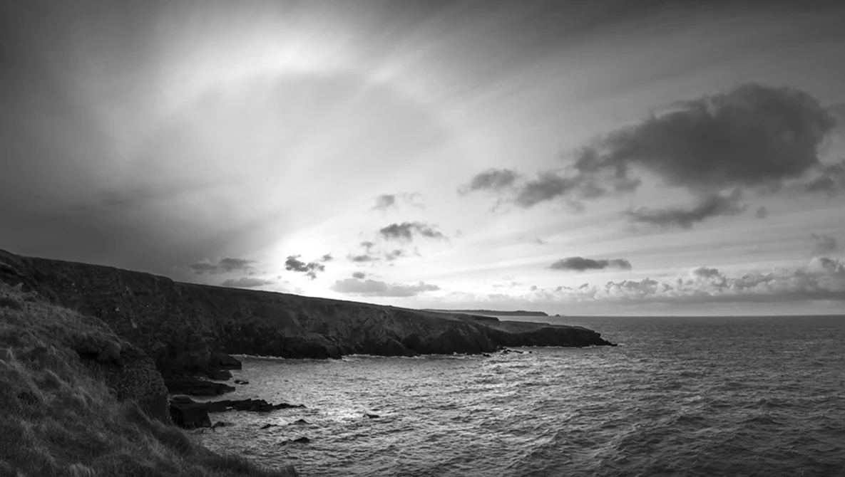 Dramatic black and white coastal landscape with rocky cliffs overlooking turbulent sea under stormy sky.