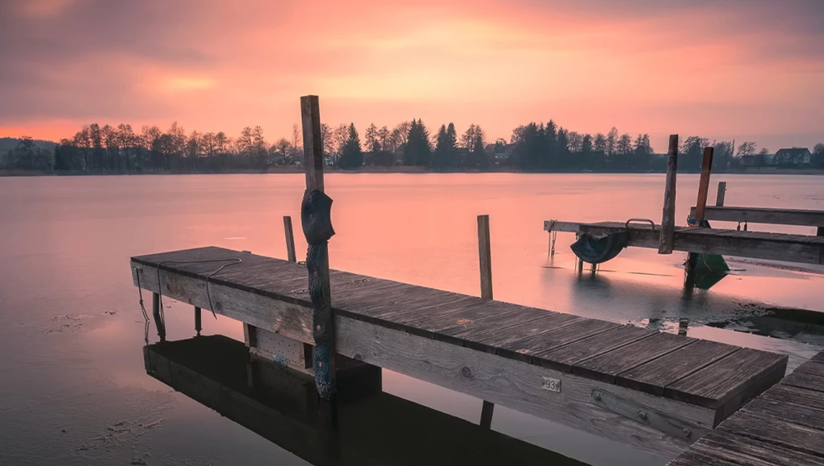 Wooden boat docks extending into a calm lake at sunrise, with pink sky and forested shoreline.