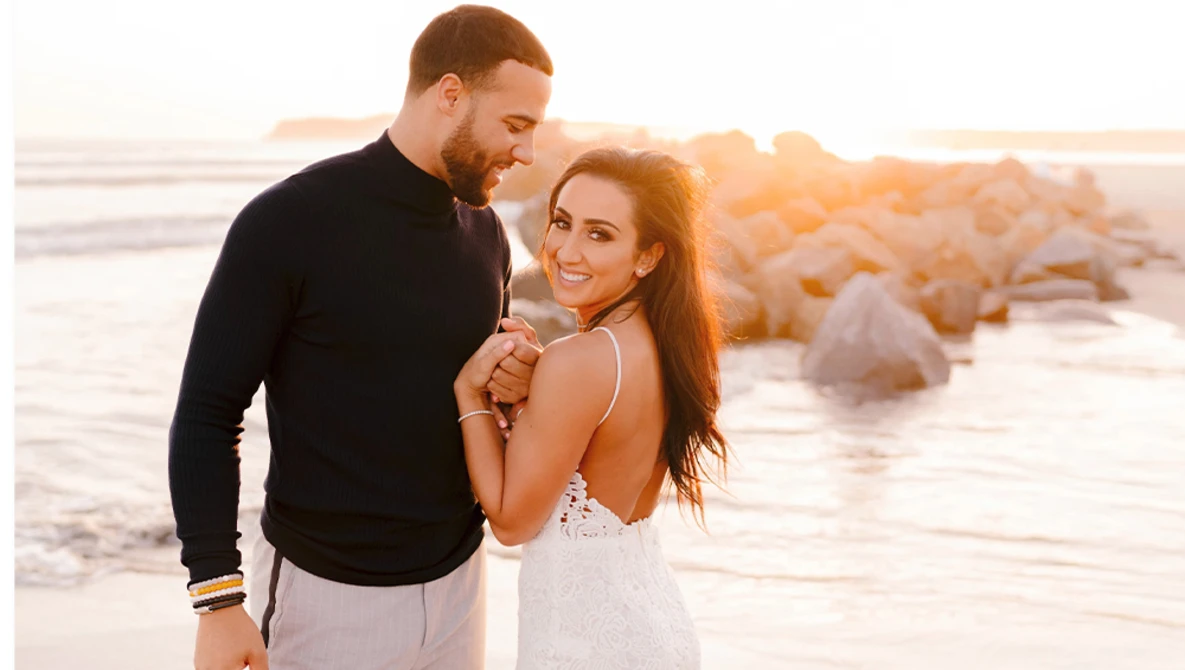 Couple smiling together on a beach at golden hour with rocky formations in the background.