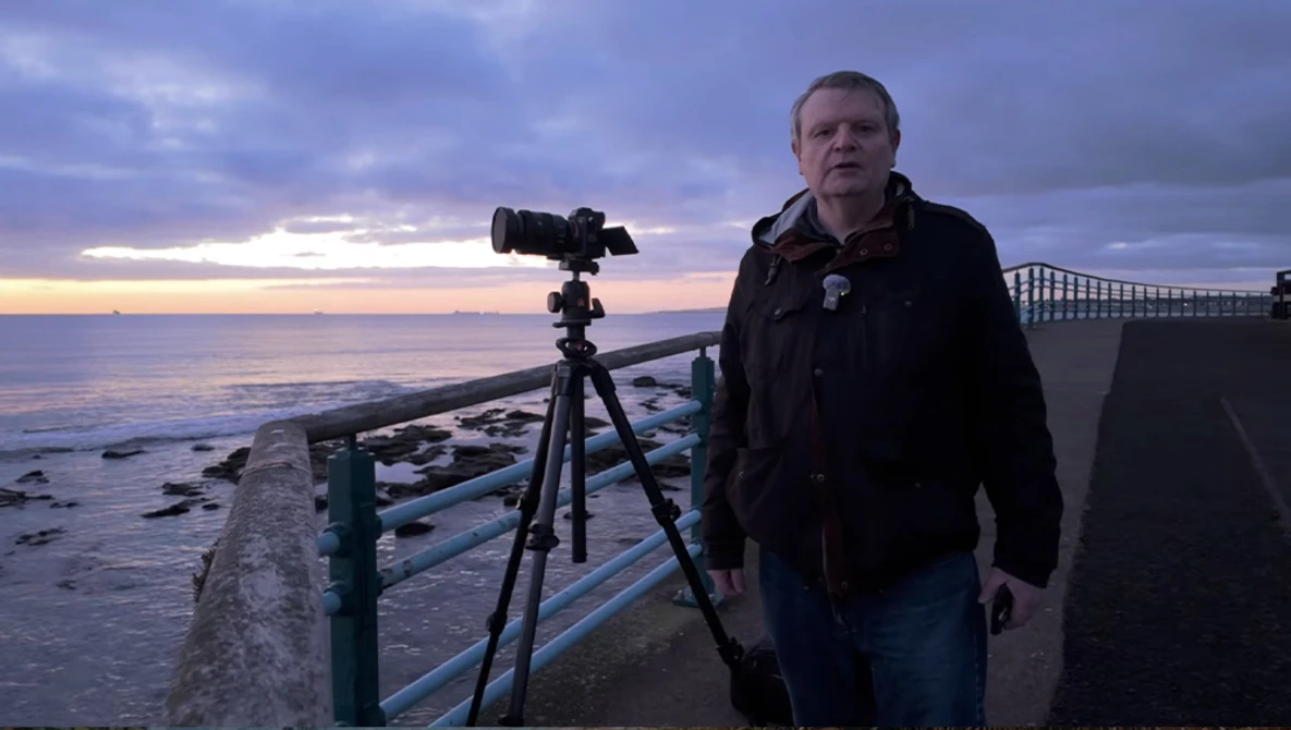 Photographer with camera on tripod at coastal location during twilight.