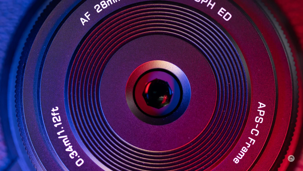 Close-up of a camera lens front element with concentric focus rings and text markings, illuminated by blue and red lighting.