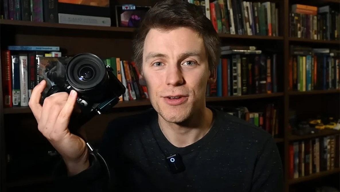 Man holding a mirrorless camera with a prime lens, speaking toward the camera in a home studio setting.
