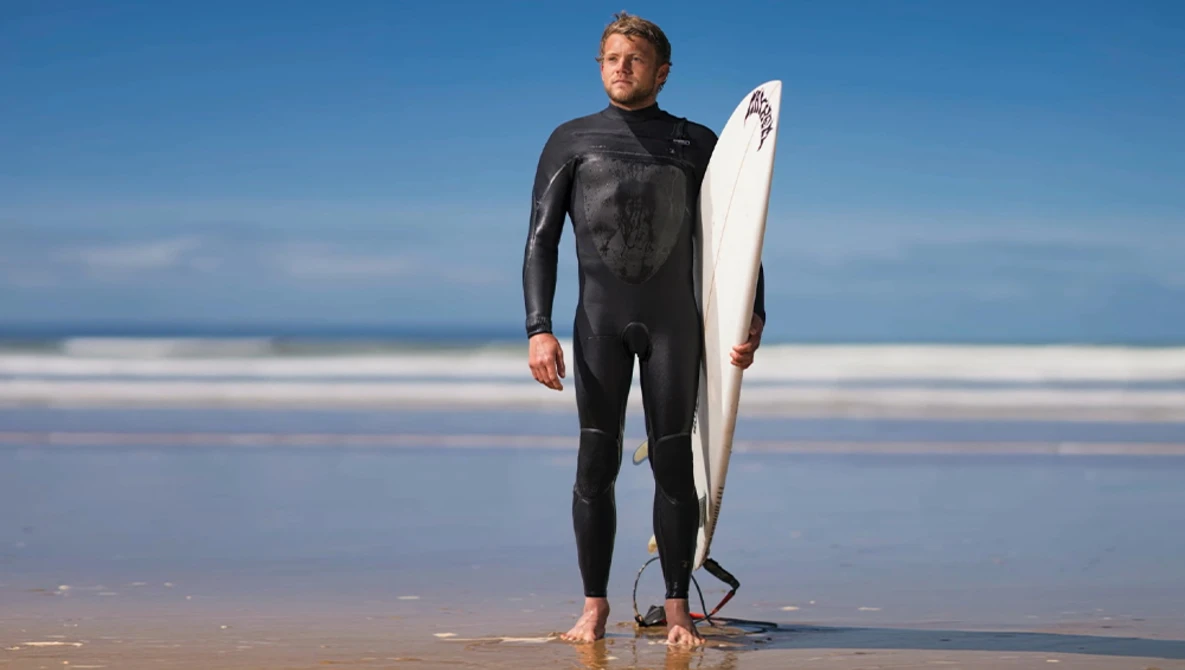 Surfer in black wetsuit holding white surfboard on wet sand beach at golden hour.