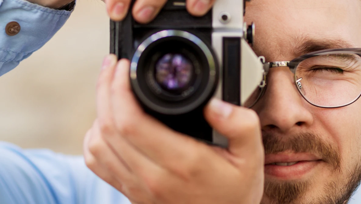 Photographer wearing glasses peers through rangefinder camera viewfinder while holding the camera up to his eye.