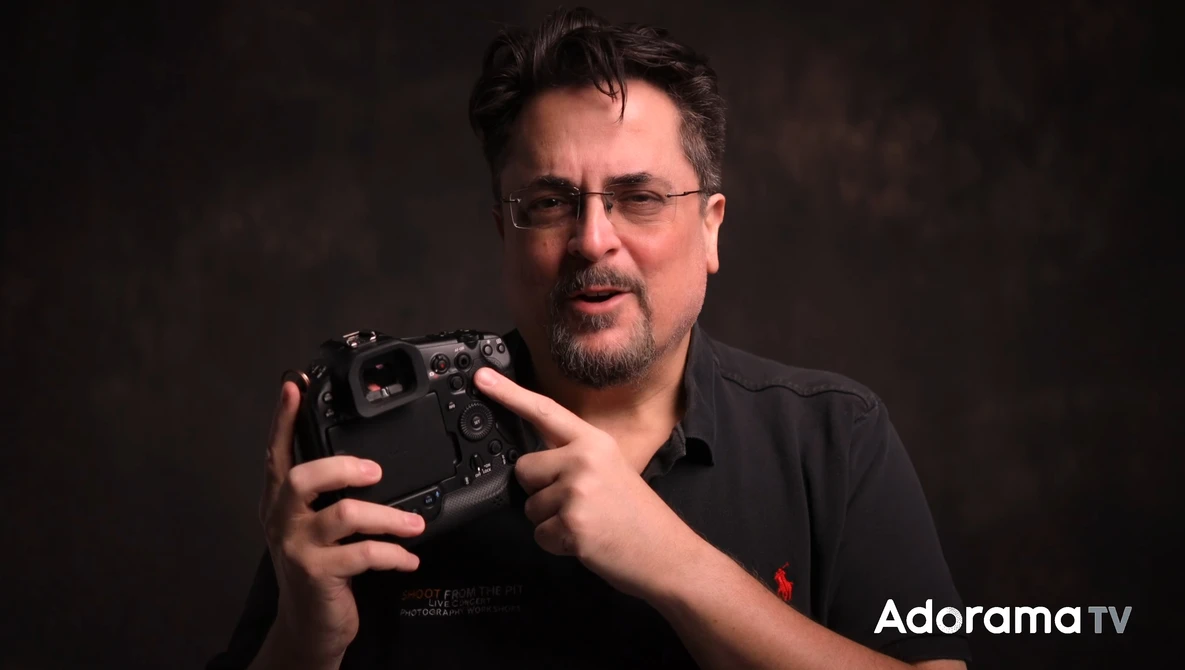 Man with glasses holding a camera controller against a dark background.