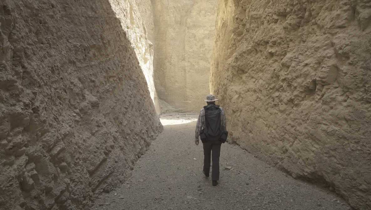 Lone figure walking away through a narrow alleyway between high earthen or mud-brick walls.