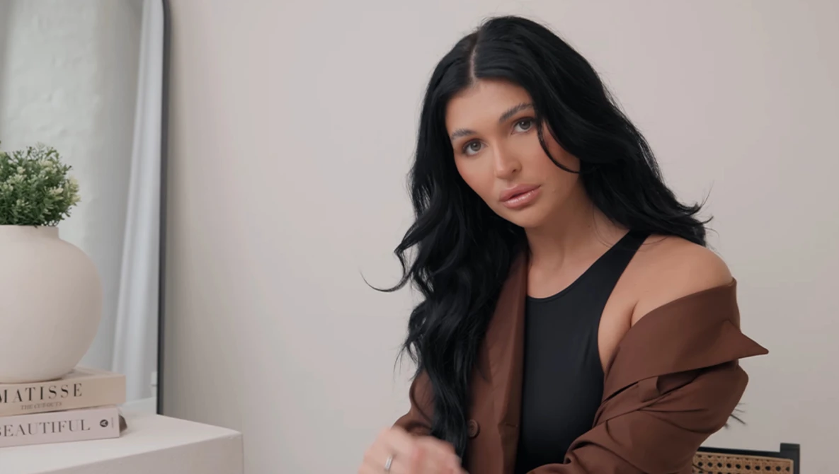 Woman with long black hair wearing a brown and black layered jacket, photographed indoors against a neutral wall.
