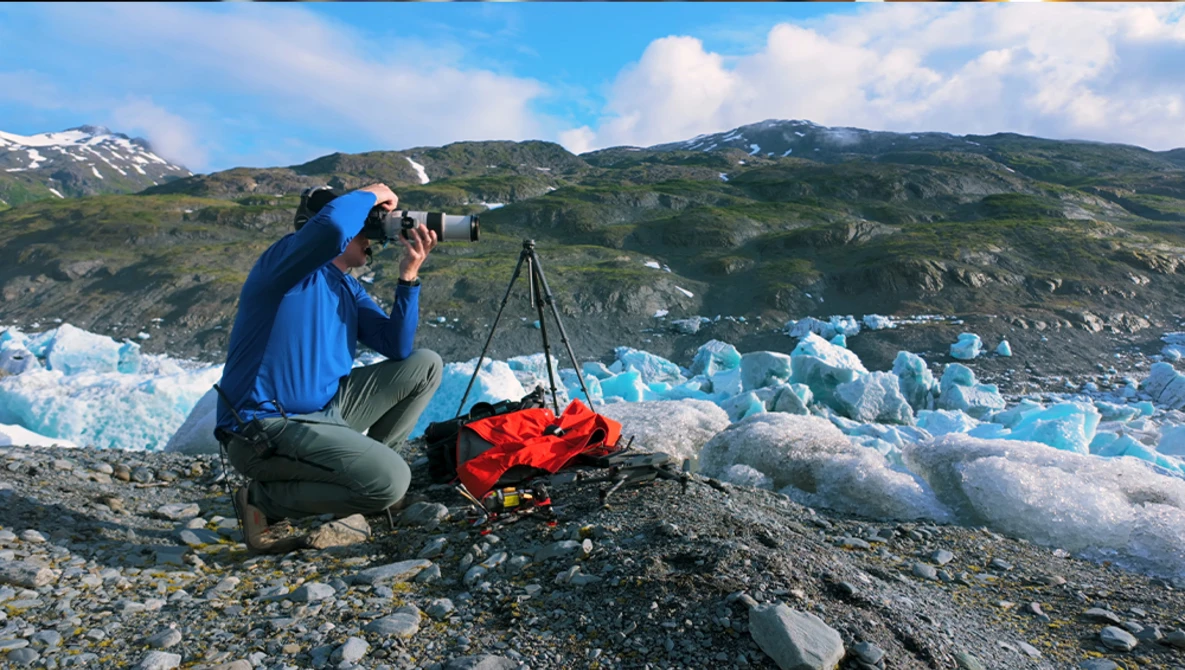 Photographer crouching with camera on tripod, documenting blue glacier icebergs with mountains in background.