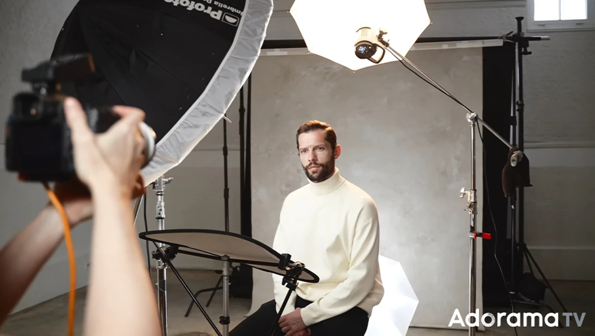 Photographer operating camera while subject sits during studio portrait session with professional lighting setup.