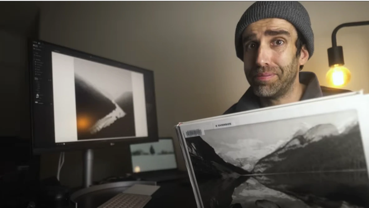 Photographer holding a landscape print while reviewing images on dual monitors in a studio workspace.