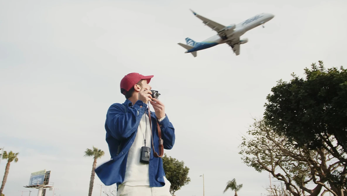 Man in blue jacket and maroon cap watches airplane flying overhead while holding binoculars to his face.